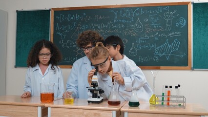 Skilled diverse children looking under microscope while caucasian student prepare chemical solution. Professional teacher looking after student doing experiment while sitting at blackboard. Pedagogy.