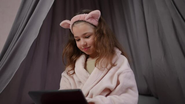 Slow motion. A girl in a bathrobe with a pink cosmetic hairband on her head looks carefully at the tablet screen, smiles and talks. A little girl sits in a beautiful bedroom and communicates using a