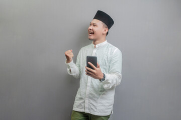 Asian Muslim man clenching his fist and holding his smartphone while looking at an empty space