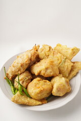 A plate of assorted fried snacks including stuffed tofu, tempeh, and fritters served with bird's eye chili. Captured against a white background. Popular for breaking fast and snacking.