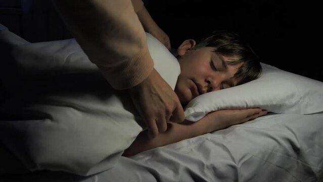 The hands of a caring young mother cover the shoulders of her sleeping son using a warm blanket, caring and loving the child. Healthy sleep eleven year old teenage boy in bed with white linen.