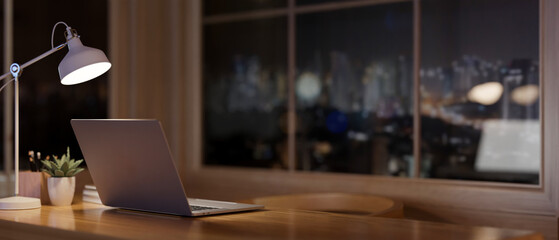A laptop on a desk in a contemporary room at night, with a view of the city skyscrapers at night.
