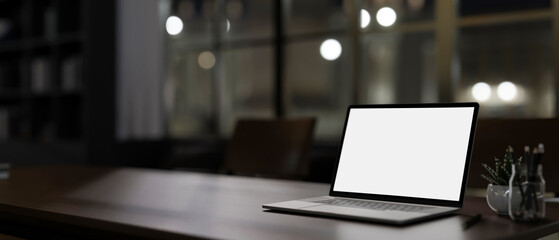 A modern dark office at night features a white-screen laptop computer mockup on a table.
