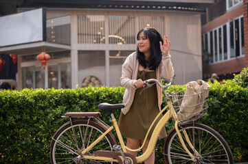 A friendly Asian woman waves her hand to say hi to her friend while pushing her bike in the city.
