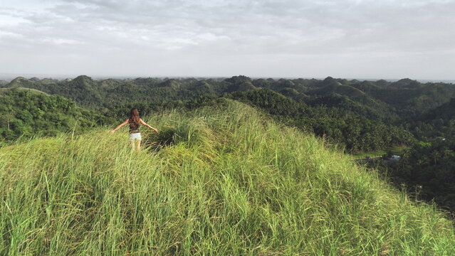 Woman rising up hands aerial view: green grass mount top in tropical Philippines. Traveler girl on hill grassy peak fill freedom of wild asian nature. Epic landcape of exotic mountains in drone shot
