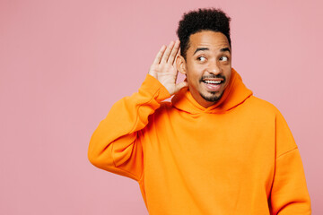 Young curious nosy man of African American ethnicity wear yellow hoody casual clothes try to hear you overhear listen intently isolated on plain pastel light pink background studio. Lifestyle concept.