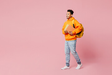 Full body young smart student man of African American ethnicity in yellow hoody casual clothes backpack bag hold books walk go isolated on plain pastel pink background High school university concept