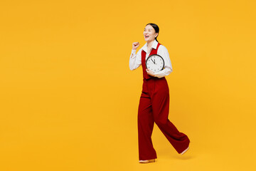 Full body side view young lawyer employee business woman of Asian ethnicity in formal red vest shirt work at office hold in hand clock run do winner gesture isolated on plain yellow background studio.