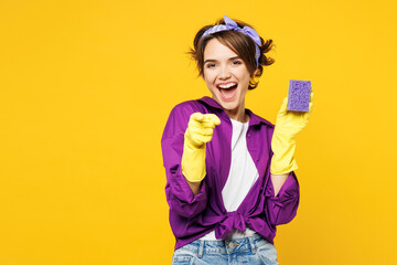 Young happy woman wear purple shirt rubber gloves do housework tidy up hold in hand sponge point index finger camera on you isolated on plain yellow background studio portrait. Housekeeping concept.