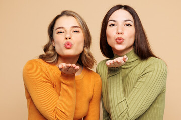 Young lovely friends two women they wear orange green shirt casual clothes together blow send air kiss look camera isolated on plain pastel light beige background studio portrait. Lifestyle concept.