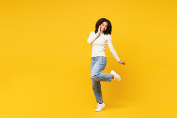 Full body little kid teen girl of African American ethnicity wear white casual clothes stand hold put hand on face raise up leg isolated on plain yellow background studio. Childhood lifestyle concept. - 763828707