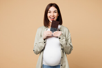 Young happy pregnant expectant woman future mom wear grey shirt with belly stomach tummy with baby hold eat bite bar of chocolate isolated on plain beige background Maternity family pregnancy concept