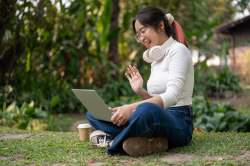 A positive, happy Asian woman is having an online meeting in a green park, sitting on the grass.