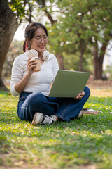 A happy young Asian woman working remotely in a green park on a bright day, using her laptop.