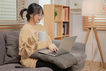 An attractive Asian woman is working remotely at home, sipping coffee, and working on her laptop.