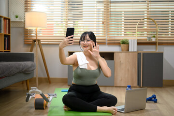 Pretty young woman sitting on exercise mat and making video call over mobile phone at home