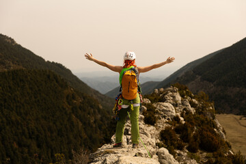 A woman is standing on a mountain top with a backpack on her back. She is wearing a helmet and she...