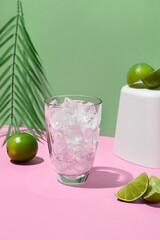 Side view of a transparent cocktail glass with ice on a pink background, green backdrop, white podium with lime. Minimalist, modern style for a refreshing, eye-catching drink. Vertical composition