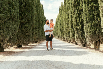 a father carries his son amidst a perfect avenue lined with cypress trees, Travelling with kids.