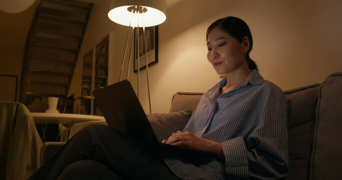 A woman puts on headphones and typing on a laptop sitting on the couch in an apartment, dimmed lights. She smiles