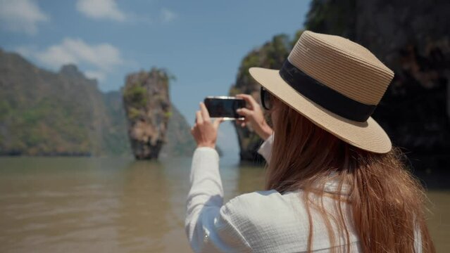 Woman travels on island Khao Phing Kan. Young female tourist sits on a boat and films take a photo of beautiful mountain landscape with a rock in the sea on smartphone. Travel, tourism concept.