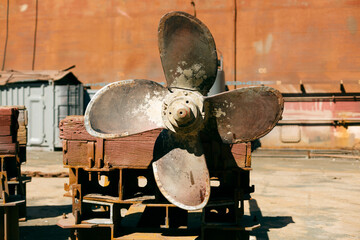 Large, rusted ship propeller displayed on wooden blocks in a shipping yard, symbolizing industrial...