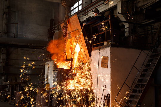 Industrial worker oversees molten metal pouring from furnace at steel mill. Foundry specialist in protective gear supervises smelting process. Heavy industry, high temp steel production. - Powered by Adobe