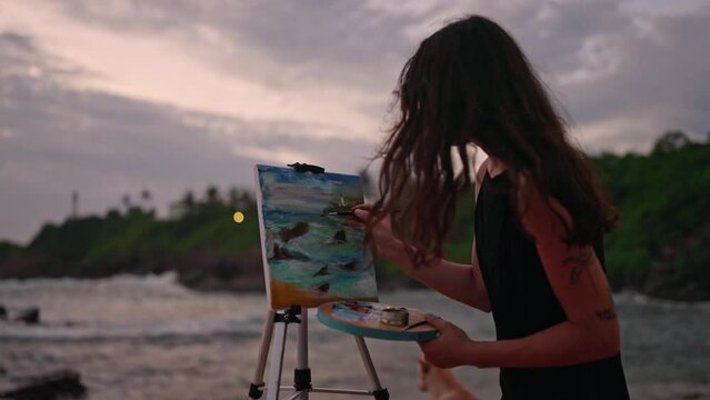 Female artist paints tumultuous sea on canvas at twilight on sandy shore. Waves crash, moody skies above, paintbrush in hand, focused on creating stormy marine artwork, inspiration flowing. Slowmo