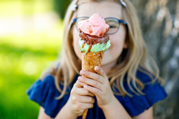 Happy preschool girl eating colorful ice cream in waffle cone on sunny summer day. Little toddler...