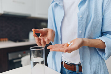 Woman holding pills and glass of water standing in front of kitchen counter, taking medication for health and wellness