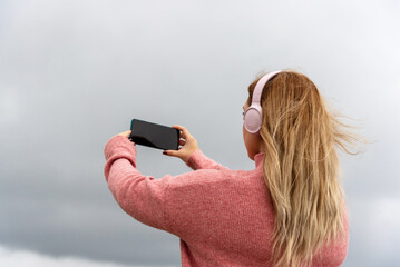 Long-haired blonde woman with her back turned, wearing a pink jumper and large matching headphones, listening to music by the sea and taking a picture of the landscape on a cloudy day.
