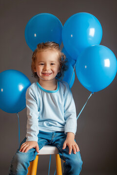 Studio portrait of a lovely smiling child with blue balloons  sitting on a stool, cute little boy or girl, birthday party, school photograph happy festive celebration or autism awareness day symbol