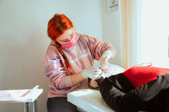 Woman piercer in a mask and gloves makes a piercing of a girl&rsquo;s ears on a couch by the window in a salon