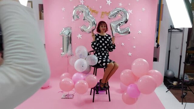 A photographer photographs a beautiful girl in a polka dot dress on a pink background on her birthday. Photo session in the studio