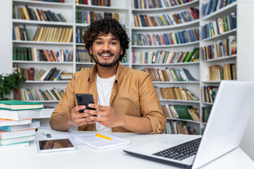 A cheerful young man with curly hair smiles while studying from home, surrounded by books, a...