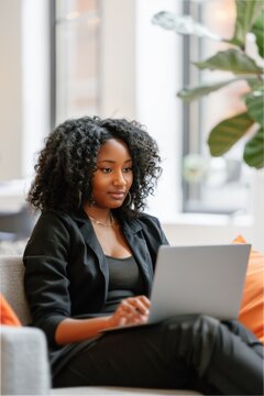 young african american business woman using laptop sitting on a couch in the hotel lobby with green plants. busy professional black female entrepreneur holding computer on lap working