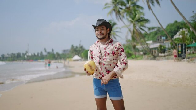 Confident man strolls sandy shore, floral shirt, cowboy hat, holds coconut drink. Smiling, enjoys tropical beach, sunny day. Fashionable gay traveler, diverse, vibrant LGBT vacation scene.