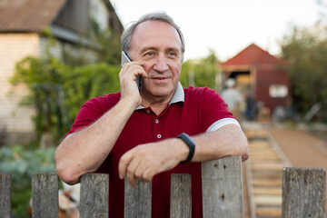Mature man standing relaxed near fence talking on phone in backyard smiling