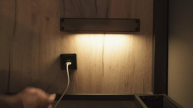 Tourist plugs USB cable into built in USB socket in hotel under night lamp on wall. Closeup of man charging discharged electronic gadgets. Convenience and modern service in apartments.