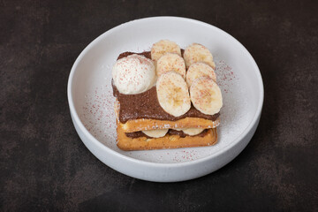 Belgian waffles with banana and chocolate sauce on dark background angle view.