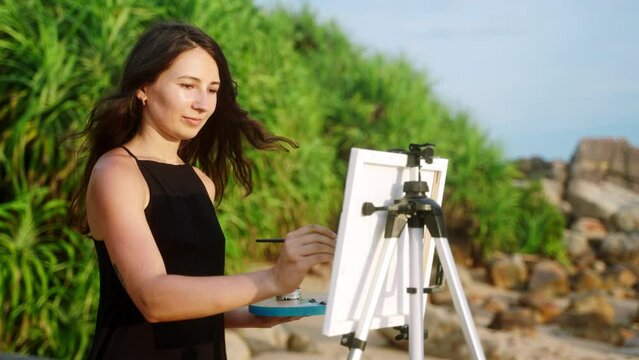 Artistic woman paints seascape on beach easel. Creative outdoor painting session by ocean. Brunette artist with brush captures coastal view on canvas. Hobbyist in black dress enjoying art, nature.