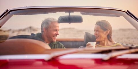 Retired Senior Couple In Classic Sports Car With Hot Drink At Beach Watching Sunrise