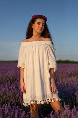 Young brunette girl looking at camera, in white dresse posing in lavender  field, beautiful sunset background. Vertical view.