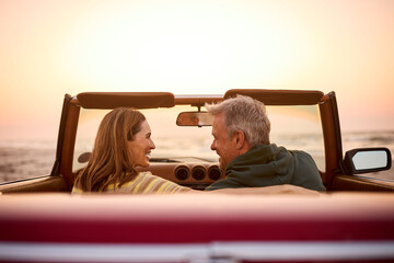 Rear View Of Retired Senior Couple On Vacation In Classic Sports Car At Beach Watching Sunrise