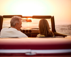 Rear View Of Retired Senior Couple On Vacation In Classic Sports Car At Beach Watching Sunrise - Powered by Adobe