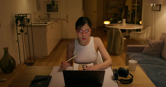 A woman with glasses, dressed in casual clothes, eats rolls with chopsticks sitting in front of an open laptop at home, dim evening light