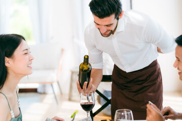 A man in a white shirt is pouring wine for a woman and a man at a table. The woman is wearing a green dress. The table is set with wine glasses, happy love couple dating in dinner time