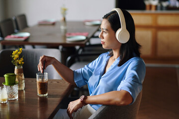 Pensive woman listening to music in headphones and drinking iced coffee in cafe