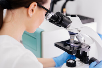 In a modern medical laboratory: a microbiologist in glasses looks under a microscope, analyzing a sample. Scientist working with high-tech equipment. Close-up