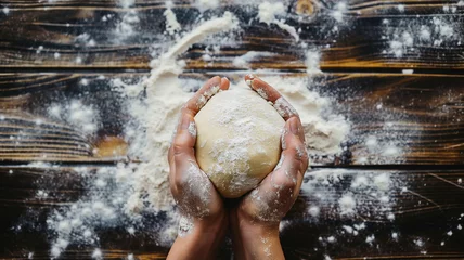 Selbstklebende Fototapeten Hands holding fresh dough on a wooden surface. A person kneads dough, sculpts a form of bread on a wooden table with flour. Pastries, bread making, home baking process. Top view, copy space. © Mariia Mazaeva